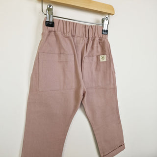 Back view of dusty pink chino-style elasticated trousers, featuring two back pockets and an Aneby hem tag on a wooden hanger on a neutral background. 