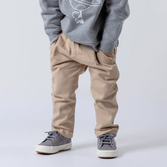 Waist-down view of a young child wearing oat-coloured chino-style elasticated trousers, standing with hands in pockets. The child is wearing a space-themed jumper against a neutral background.