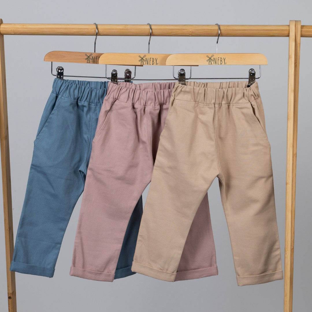 Three pairs of kids' chino-style elasticated trousers in dusty pink, slate blue, and oat, shown from the back with Aneby hem tags visible, hanging from a wooden clothing frame with Aneby-branded wooden hangers.