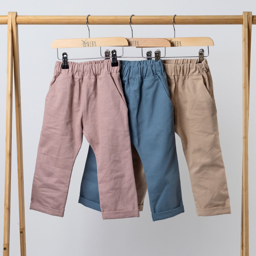 Three pairs of kids' chino-style elasticated trousers in dusty pink, slate blue, and oat, hanging from a wooden clothing frame with Aneby-branded wooden hangers.