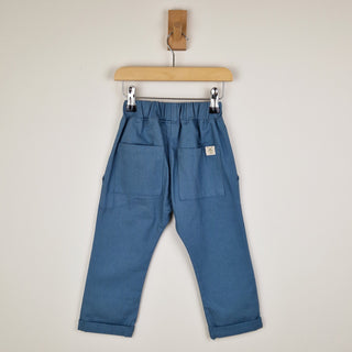 Back view of slate blue chino-style elasticated trousers, featuring two back pockets and an Aneby hem tag, hanging from a wooden hanger.