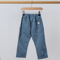 Back view of slate blue chino-style elasticated trousers, featuring two back pockets and an Aneby hem tag, hanging from a wooden clothing frame.