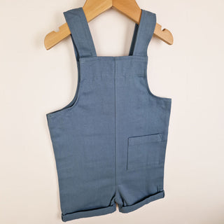Slate blue kids' cotton dungaree shorts, back view. Features a minimalist style with a rear pocket and crossover straps for a snug fit.