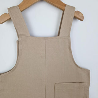 Close up of back of oat-coloured kids' cotton dungaree shorts. Detailing the back patch pocket with reinforced corner stitching and the binding stitching around the armhole and back neckline. 