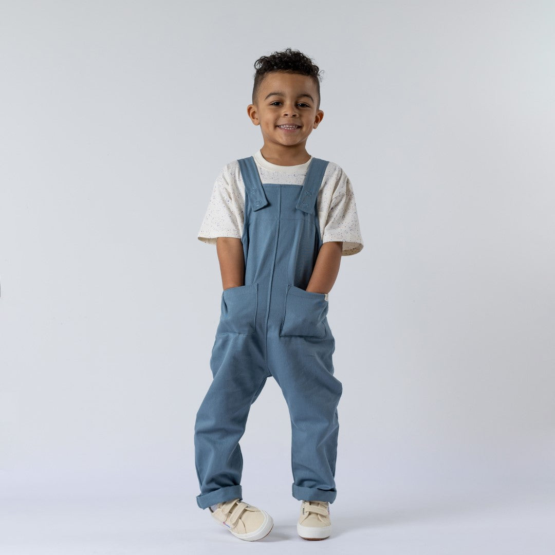 A boy wearing kids dungarees with patch pockets in blue twill. He is shown in a relaxed, natural pose against a plain white background. The image highlights the durable fabric, clear patch pocket details, and precise stitching, with natural lighting that accentuates the garment’s texture.