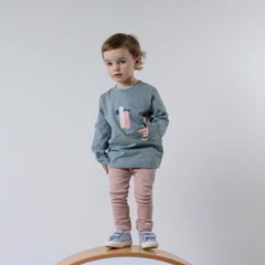 A small child wearing a kids' long sleeve artist t-shirt with a pink, blue, and oat brushstroke design, paired with dusty pink Aneby leggings. The child stands on a wooden wobble board, showcasing the relaxed fit and artistic style.