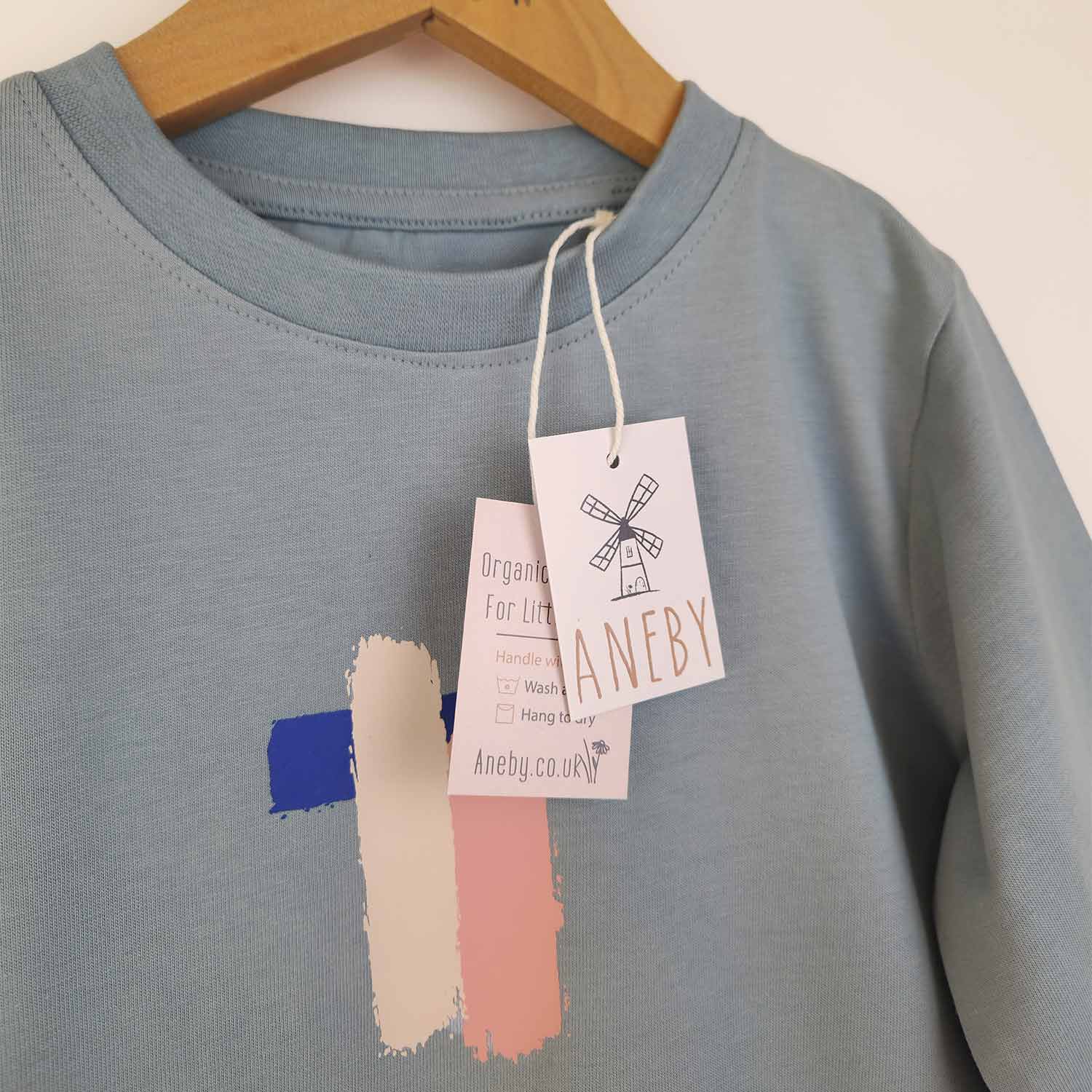Close up image of a kids' long sleeve artist t-shirt with a brushstroke pattern in pink, blue, and white. The image also shows the Aneby swing tag with was h care instructions. 
