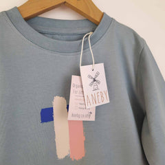 Close up image of a kids' long sleeve artist t-shirt with a brushstroke pattern in pink, blue, and white. The image also shows the Aneby swing tag with was h care instructions. 