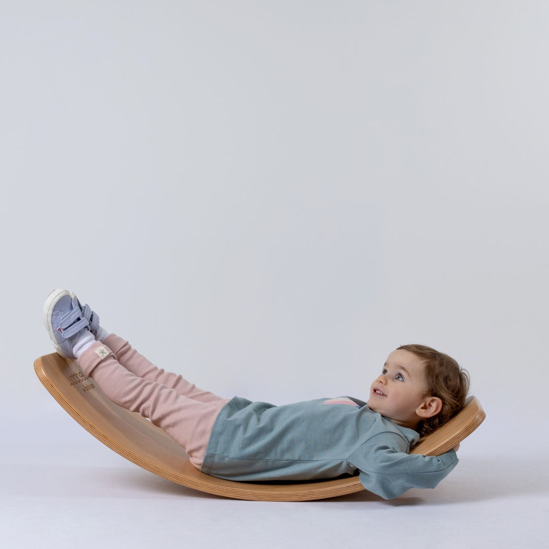 A small child wearing a kids' long sleeve artist t-shirt featuring an abstract brushstroke pattern in pink, blue, and white. Lying on a wobble board with toes pointed up, the child looks at the ceiling with a joyful grin.