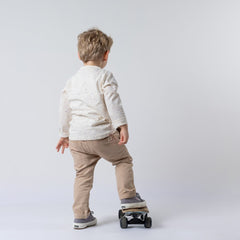 Back view of a young boy wearing a kids long sleeve ecru t-shirt and oat chinos, balancing with one foot on a skateboard against a neutral backdrop.