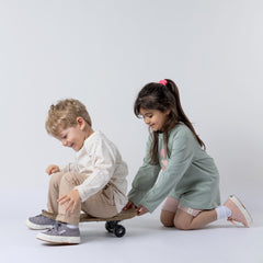Boy wearing a kids long sleeve ecru t-shirt and oat chinos, sitting on a skateboard while a girl pushes him, both smiling and playing together.
