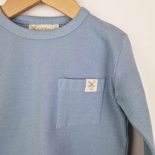 Front view of a slate blue coloured kids long sleeve ribbed t-shirt on a neutral background. Close up shot shows the details of the ribbed neck binding and the front chest patch pocket with Aneby windmill hemtag.