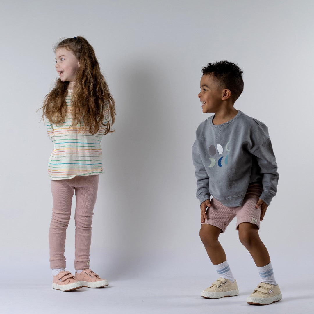 Two small children smiling and looking into the distance. The boy wears a grey kids' moon sweatshirt with six moons and dusty pink shorts. The girl wears a green striped long-sleeve t-shirt with dusty pink leggings.