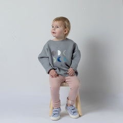 Small blonde girl sitting on a wooden stool, wearing a grey kids' moon sweatshirt with six moons on the front and dusty pink leggings. She smiles while surrounded by bubbles, looking into the distance.