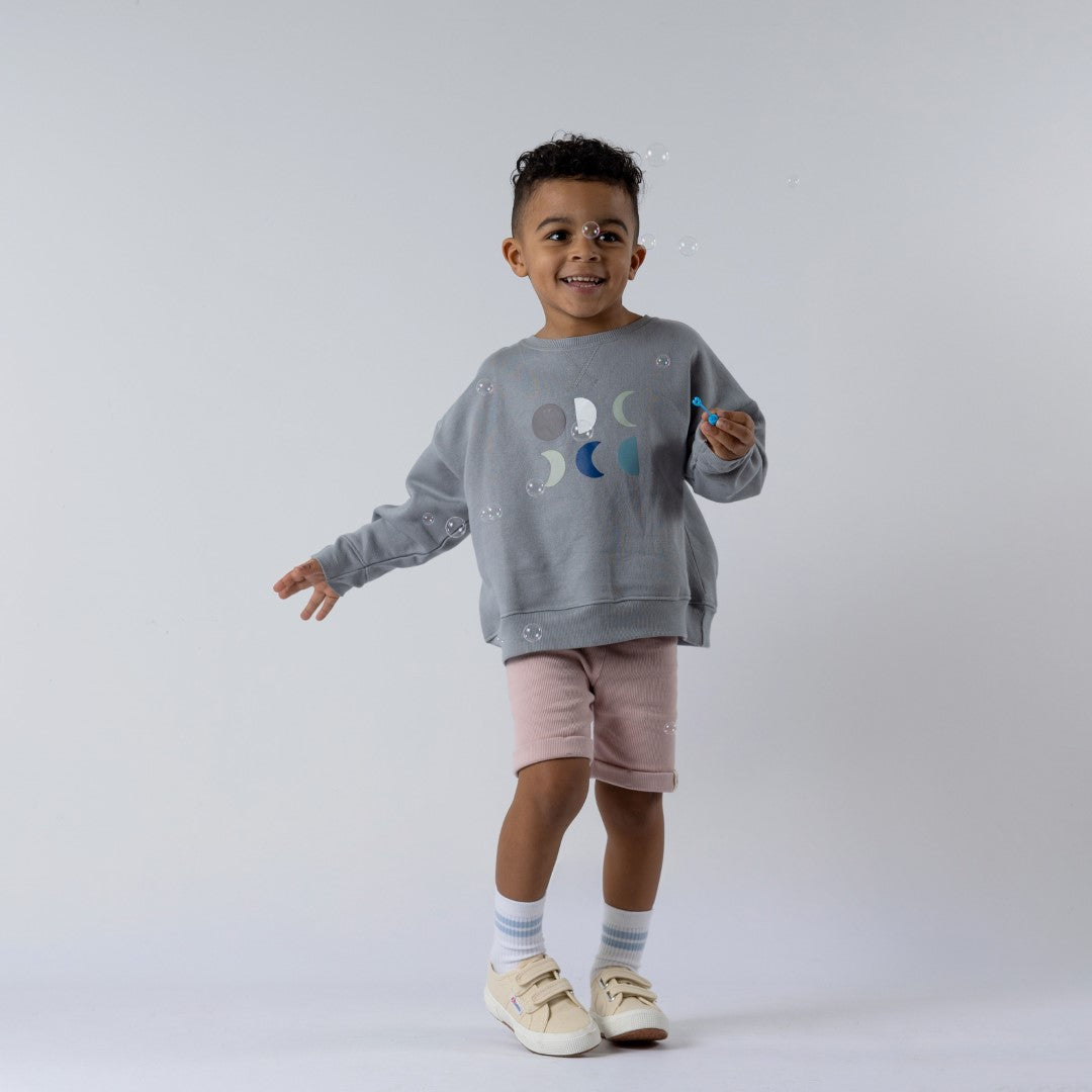 Child wearing a grey kids moon sweatshirt featuring six moons on the front, paired with dusty pink shorts. Smiling while holding a bubble wand, surrounded by bubbles, neutral background.