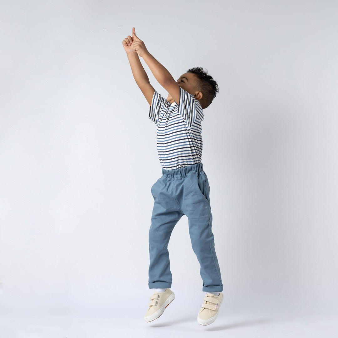 A young boy wearing a navy blue striped t-shirt with white horizontal stripes, jumping in the air while pointing to the sky. He is wearing slate blue chinos, and his joyful movement adds a sense of energy to the scene.