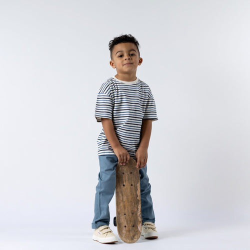 A young boy wearing a navy blue striped t-shirt with horizontal white stripes, standing sideways. He is holding a skateboard in one hand and wearing slate blue chinos. The boy’s casual stance and skateboard highlight an active, everyday look.