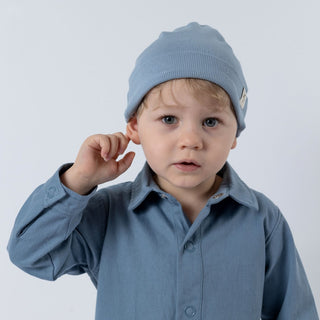 Young child wearing a soft slate blue kids ribbed cotton beanie hat, paired with a matching blue boilersuit, against a white background.