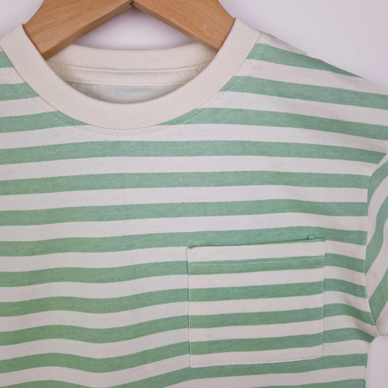 A close up photograph of the front neckline and patch pocket on a kids’ stylish green striped t-shirt, placed on a smooth surface. The t-shirt has a classic crew neckline, with a neat edhe stitch detail and a square chest patch pocket with neeat edge stitching and reinforced triangle stitch at the top corners of the pockets.