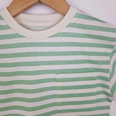 A close up photograph of the front neckline and patch pocket on a kids’ stylish green striped t-shirt, placed on a smooth surface. The t-shirt has a classic crew neckline, with a neat edhe stitch detail and a square chest patch pocket with neeat edge stitching and reinforced triangle stitch at the top corners of the pockets.