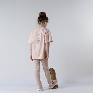 Back view of a young girl wearing a blush pink t-shirt featuring the Aneby windmill logo. She stands on a vintage wooden skateboard, dressed in pink leggings and trainers.