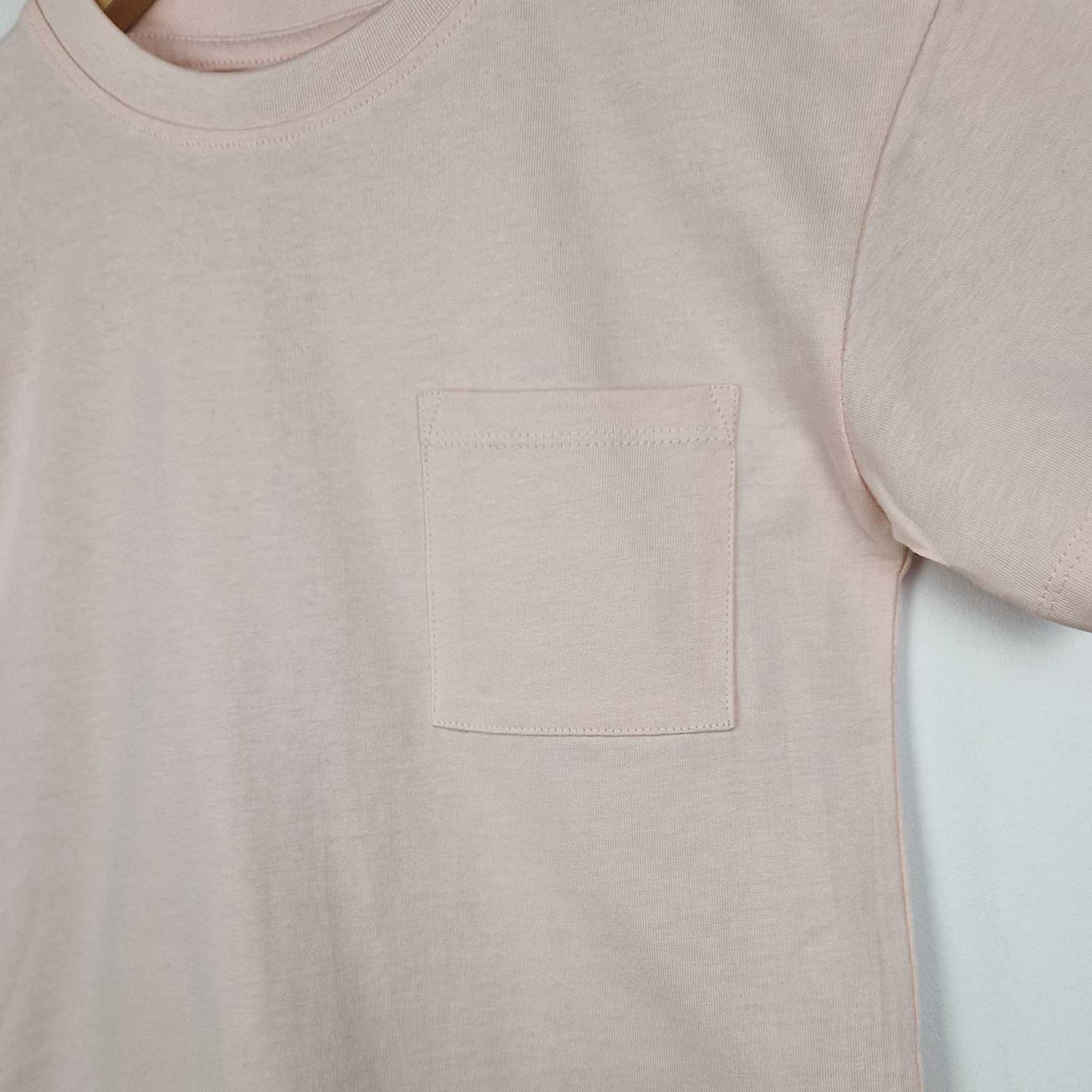 Flat lay close up image of a blush pink kids t-shirt, showing the front design which features a pocket on the left side.
