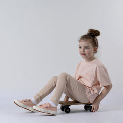 Young girl sitting on a skateboard, wearing a blush pink windmill t-shirt looking off into the distance