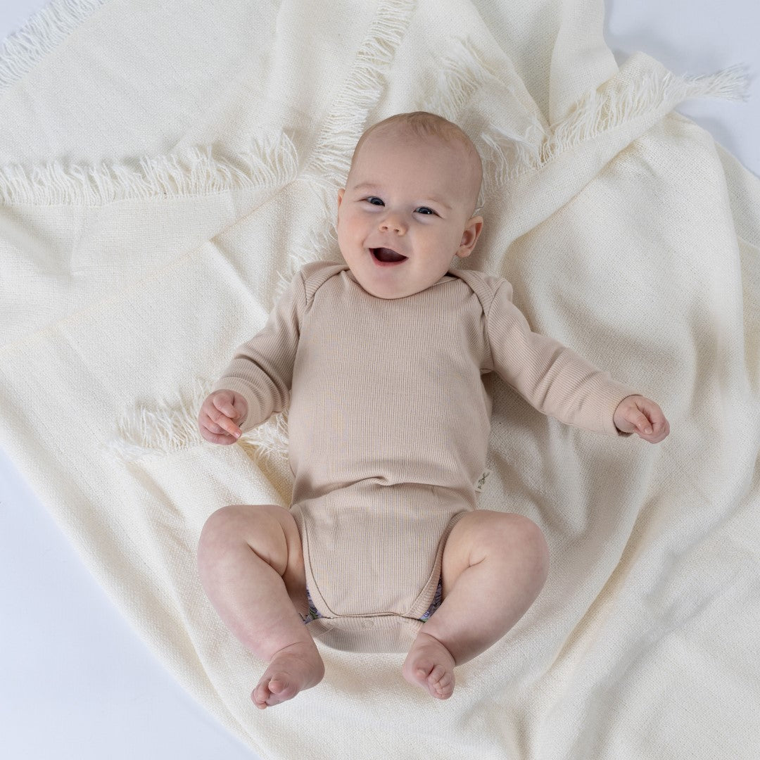 Small baby laying on a cream blanket looking straight up at the camera laughing, wearing oat-coloured ribbed bodysuit with long sleeves for babies. 