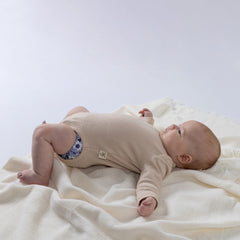 Baby wearing an oat-coloured ribbed bodysuit with long sleeves, shown from the side with the Aneby hem tag visible.