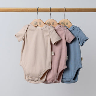 A set of three short sleeve baby bodysuits in dusty pink, slate blue, and oat colours, displayed hanging from a natural wooden rail.