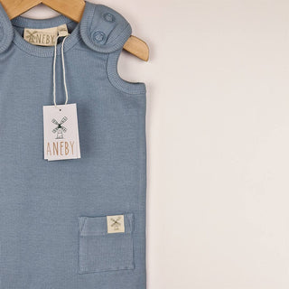 Slate blue sleeveless rib baby romper hanging on a wooden hanger, close up view showing shoulder tab with metal ring poppers and square patch pocket on a neutral background.