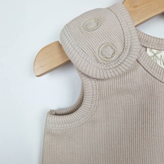 Oat-coloured sleeveless rib baby romper hanging on a wooden hanger close up view showing shoulder tab with metal ring poppers and neat binding around armhole and neckline. Neutral background.